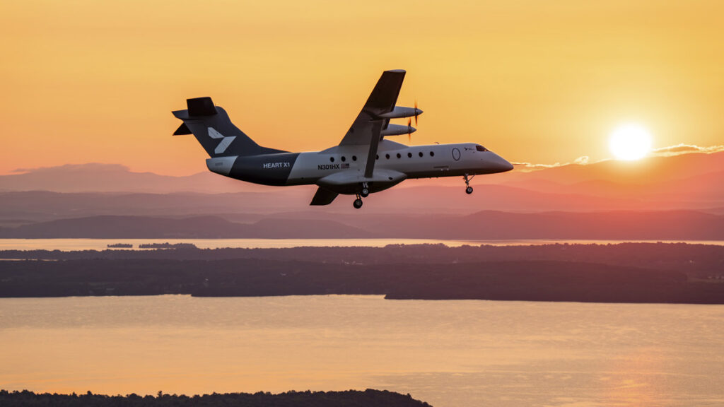 Voló el mayor avión eléctrico del mundo: promete reducir 40% los costos Avión eléctrico Heart X1 con tren de aterrizaje extendido en pleno vuelo al atardecer sobre un lago y zona montañosa.