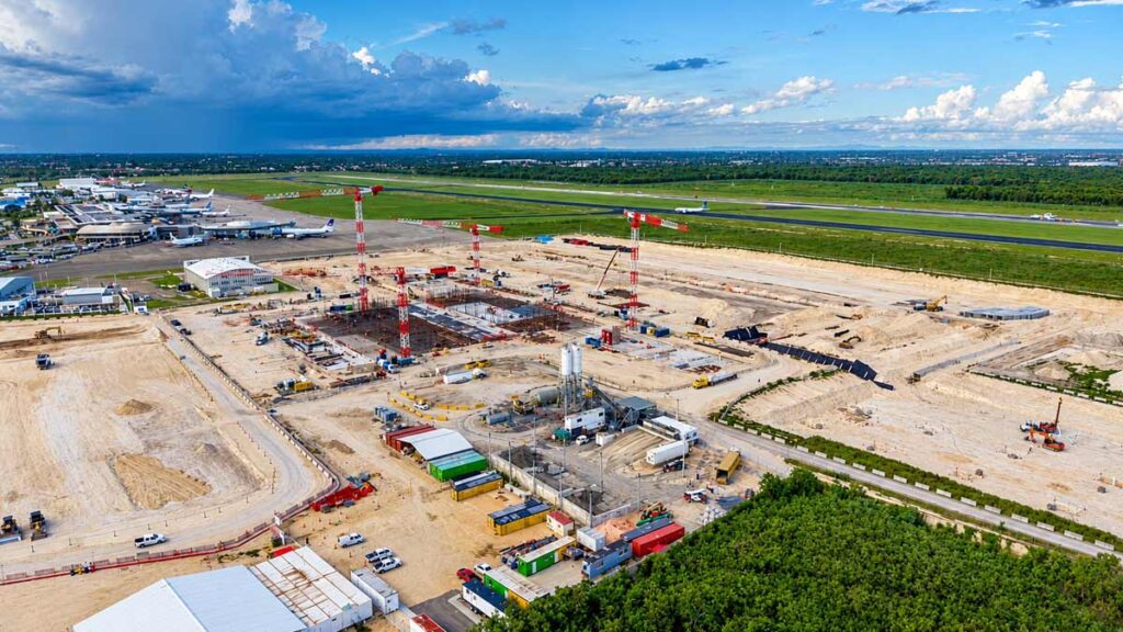 AERODOM invierte USD 400M en la nueva terminal del Aeropuerto de Las Américas Vista aérea de una obra en construcción en un aeropuerto con tres grúas torre y cimientos de hormigón junto a una pista.