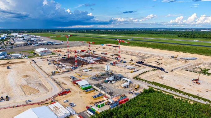 Vista aérea de una obra en construcción en un aeropuerto con tres grúas torre y cimientos de hormigón junto a una pista.